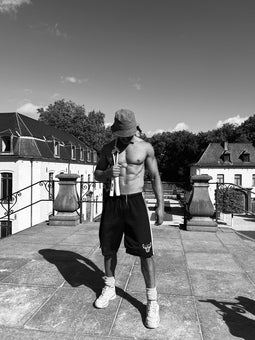 Homme torse nu en tenue sportive sur une plateforme extérieure, tenant une corde à sauter lestée par le cordon, les poignées pendantes. La corde repose sur son épaule droite. Ciel bleu en arrière-plan, l'homme porte un bob.