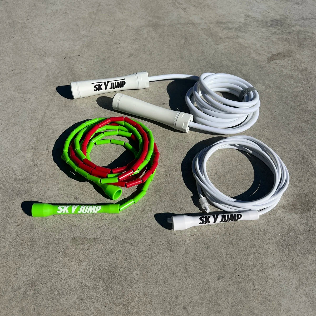 Trio de cordes à sauter SkyJump : une corde lestée blanche 500 g, une corde perlée multicolore rouge et verte (style Maroc) et une corde PVC blanche, posées sur un sol en béton clair. »